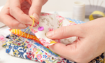 Close up of a person sewing on a purple button on  floral fabric holding a needle with yellow thread